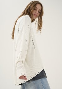 Oversized witte gebreide trui met versleten details, geribde boorden en zoom. Gecombineerd met lichtblauwe spijkerbroek.