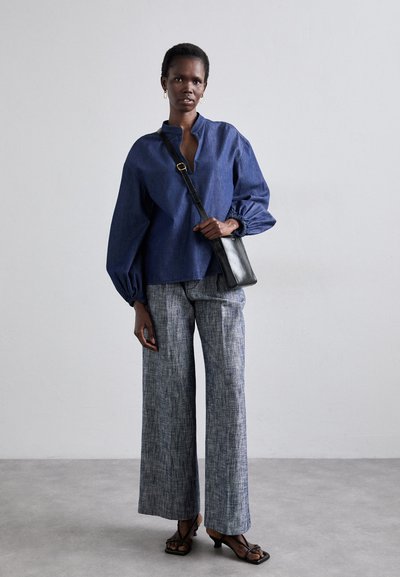 Blaue Denim-Tunika mit gepufften Ärmeln, kombiniert mit weit geschnittenen, strukturierten grauen Hosen und einer schwarzen Schultertasche. Dazu werden schwarze Absatzsandalen mit Riemchen getragen.