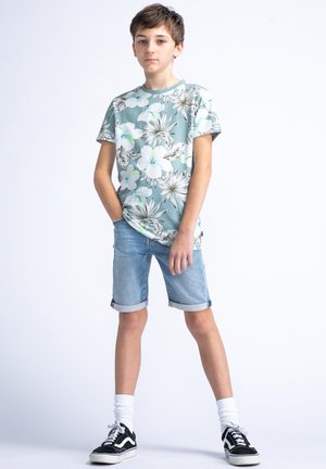 T-shirt floreale blu chiaro con maniche corte, abbinata a pantaloni corti di jeans arrotolati. Sneakers bianco e nere con calzini bianchi. Outfit casual.