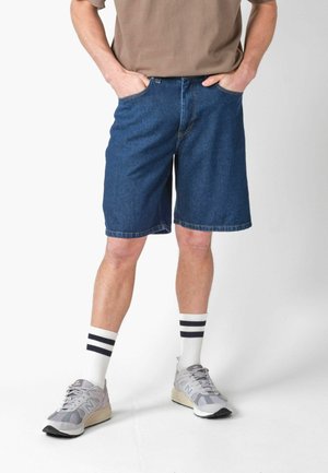 Férfi sötétkék farmer rövidnadrágban, fehér sportzokniban fekete csíkokkal, szürke futócipőben és barna pólóban, kezét zsebében tartva.