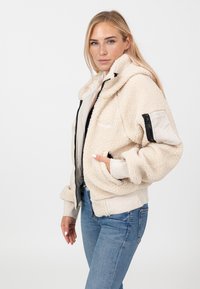 Kremowy, strukturalny polarowy dres z kapturem, ściągaczami na rękawach i bocznymi kieszeniami. Posiada zewnętrzną kieszeń z zamkiem błyskawicznym na rękawie.