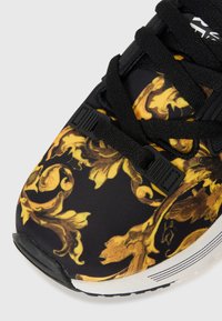 Chaussure de sport noire avec un motif floral jaune, tige en tissu texturé, lacets noirs et semelle en caoutchouc blanc avec des accents noirs.