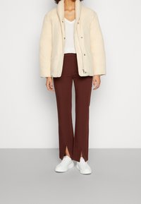 Puffig, krämfärgad jacka med hög krage, bruna raka byxor med slitsvidder och vita sneakers mot en neutral bakgrund.