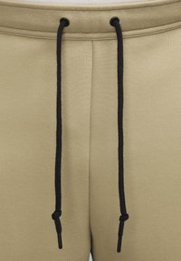 Beige joggers med en strukturerad tyg, svarta snörning och metallaglets i ändarna. Designen inkluderar ett elastiskt midjeband.