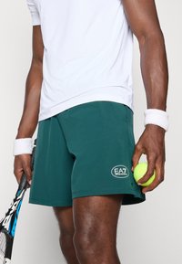 Calções atléticos verdes feitos de um tecido suave, com um patch de logo, fenda lateral, e combinados com uma raquete de ténis e uma bola.