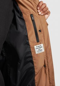 Interior de una chaqueta aislante marrón con forro negro, que presenta un bolsillo con cremallera y una etiqueta que dice: "Un abrazo cálido hecho de tela."