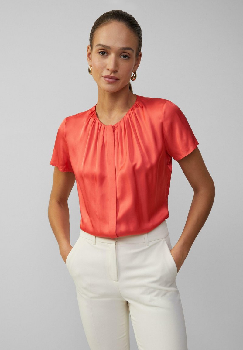Kurzärmlige, korallenfarbene Satinbluse mit rundem Ausschnitt, gefältelter Vorderseite und glatter Oberfläche, kombiniert mit maßgeschneiderten weißen Hosen.