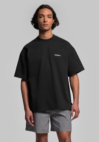 Svart bomull T-shirt med korta ärmar och rund halsringning, med vit broderad text på bröstet. Matchad med randiga shorts.