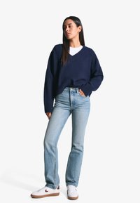 Marineblauwe gebreide trui met een V-hals en geribbelde manchetten, gecombineerd met lichtblauwe rechttoe-rechtaan jeans en witte sneakers met bruine accenten.