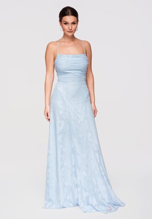 Femme aux cheveux foncés portant une robe longue bleu pâle, sans manches, avec de fines bretelles, debout devant un fond clair uni.