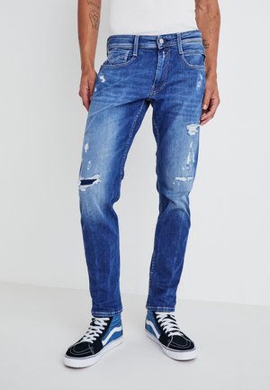 Mann trägt blaue abgenutzte Jeans und schwarze, blaue und weiße High-Top-Sneaker, steht vor schlichtem weißen Hintergrund.