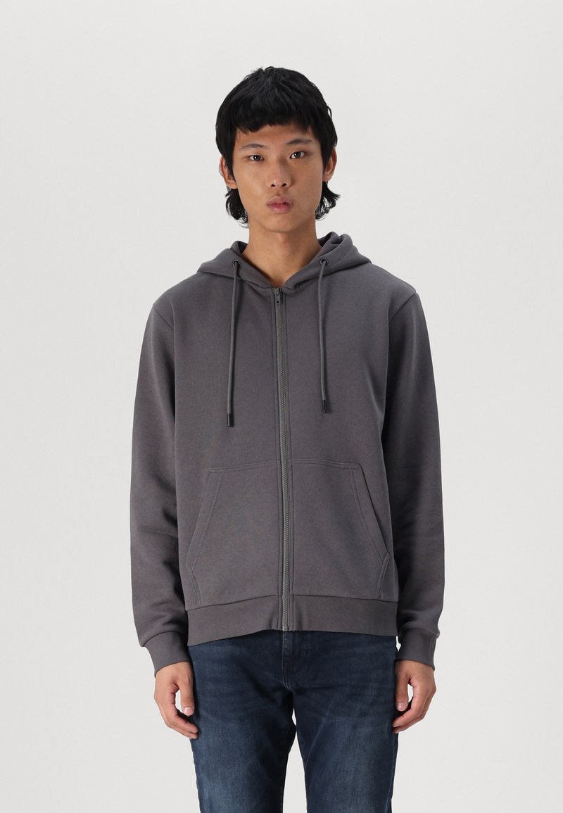 Felpa grigia con zip, realizzata in tessuto morbido, con zip frontale, cappuccio con cordino e due tasche laterali. Indossata con jeans scuri.