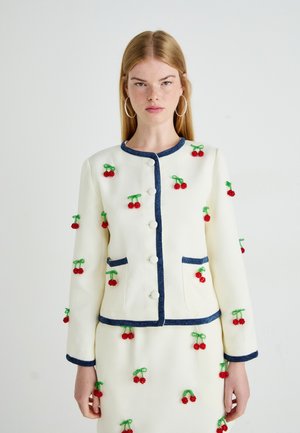 Young woman in white buttoned jacket and skirt with blue trim and red beaded cherry embellishments, standing against a plain background.