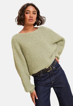 Hellgrüner, strukturierten Strickpullover mit weitem Ausschnitt und langen Ärmeln, kombiniert mit dunkelblauen Jeans und einem schwarzen Gürtel.