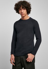 Juodas ilgas rankoves marškinys su tekstūruota medžiaga ir raglan rankovėmis, derinamas su tamsiomis kelnėmis, modelis pozavo prieš neutralų foną.