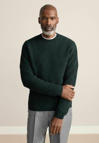 Dunkelgrüner, gerippter Strickpullover mit Rundhalsausschnitt, taillierten Ärmeln und strukturierter Oberfläche, kombiniert mit hellgrauen Hosen.