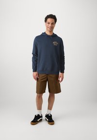 Felpa blu navy con tasca a canguro frontale, grafica dorata sul petto, abbinata a pantaloni corti marroni. Scarpe nere con accenti bianchi e calzini bianchi.