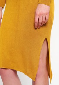 Main reposant sur une robe en tricot jaune moutarde avec une fente sur le côté, montrant une partie de la cuisse et le détail du tissu texturé.
