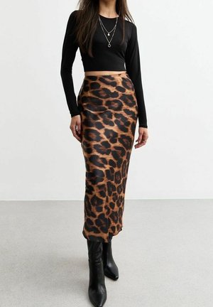 Kvinde iført sort langærmet crop top, lag-på-lag halskæder, leopardmønstret midi nederdel og sorte spidse støvler, stående på en grå gulv.