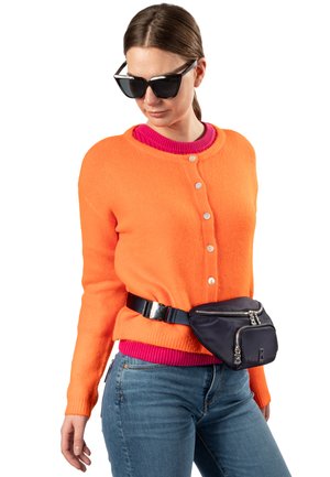 Cardigan in lana arancione con bottoni, indossato sopra un maglione rosa a coste, abbinato a jeans scuri e una borsa a tracolla nera fissata da una cintura.