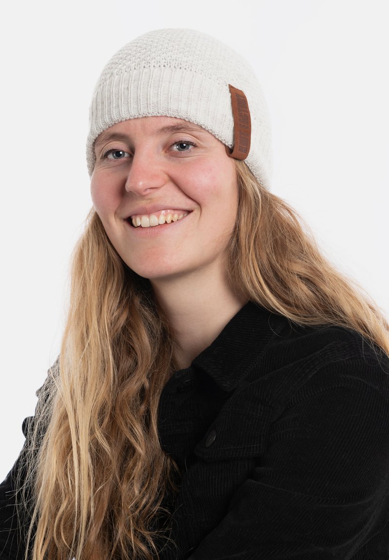 Kremowa dzianinowa czapka z efektem teksturą i brązową skórzaną łatką. Noszona z czarną kurtką z kordurowej tkaniny, z zapięciem na guziki.