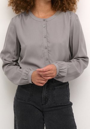 Femme portant un chemisier boutonné à manches longues gris clair avec poignets froncés et un jean noir taille haute avec des poches plaquées à l'avant.