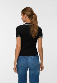 Kvinna med långt brunt hår i en hästsvans, iförd en svart kortärmad stickad topp med vit kant och blå jeans, vänd bort från kameran mot en enkel bakgrund.