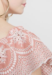 Vaaleanpunainen brodeerattu paita, jossa on monimutkaisia valkoisia kukkakuvioita, sahalaitaiset reunat ja pehmeä, tekstuurinen kangas. Suuret kultaiset renkaat korvakoru näkyvissä.