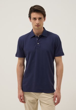 Young man standing, wearing a navy blue short-sleeve polo shirt and beige pants against a plain light background.