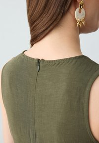 Top sans manches vert olive avec un col rond, doté d'une fermeture éclair au dos et d'une texture lisse. Les accessoires incluent de grandes boucles d'oreilles circulaires.
