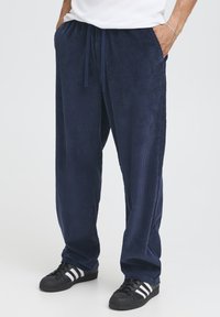 Tmavě modré manšestrové kalhoty mají volný střih, žebrovaný povrch, elastický pas se šňůrkou a boční kapsy, doplněné černými teniskami.