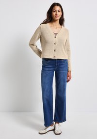 Beige Strickjacke mit V-Ausschnitt, fünf braunen Knöpfen, kombiniert mit weitem blauen Jeans und weißen Sneakers mit braunen Akzenten.