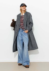Manteau en laine gris, chemise rayée bordeaux et blanche, jean large bleu clair avec des boutons sur le côté, et chaussures imprimé léopard. Tenue décontractée avec une casquette.