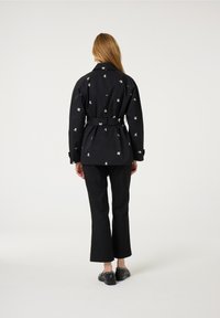 Zwarte jack met riem en wit bloemenpatroon, voorzien van een kraag en knoopmanchetten, gecombineerd met zwarte broek en gespijkerde schoenen.