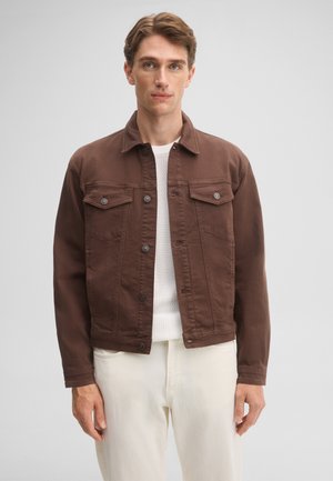 Junger Mann mit heller Haut und braunen Haaren trägt eine braune, geknöpfte Jacke über einem weißen Strickpullover und hellbeigen Hosen und steht vor einem grauen Hintergrund.