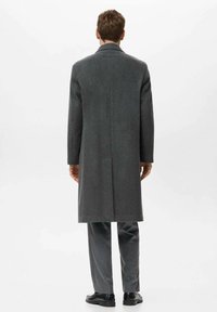 Cappotto lungo in lana grigia con taglio dritto, spacco singolo sul retro e senza chiusure visibili. Indossato sopra un pantalone grigio e scarpe nere.