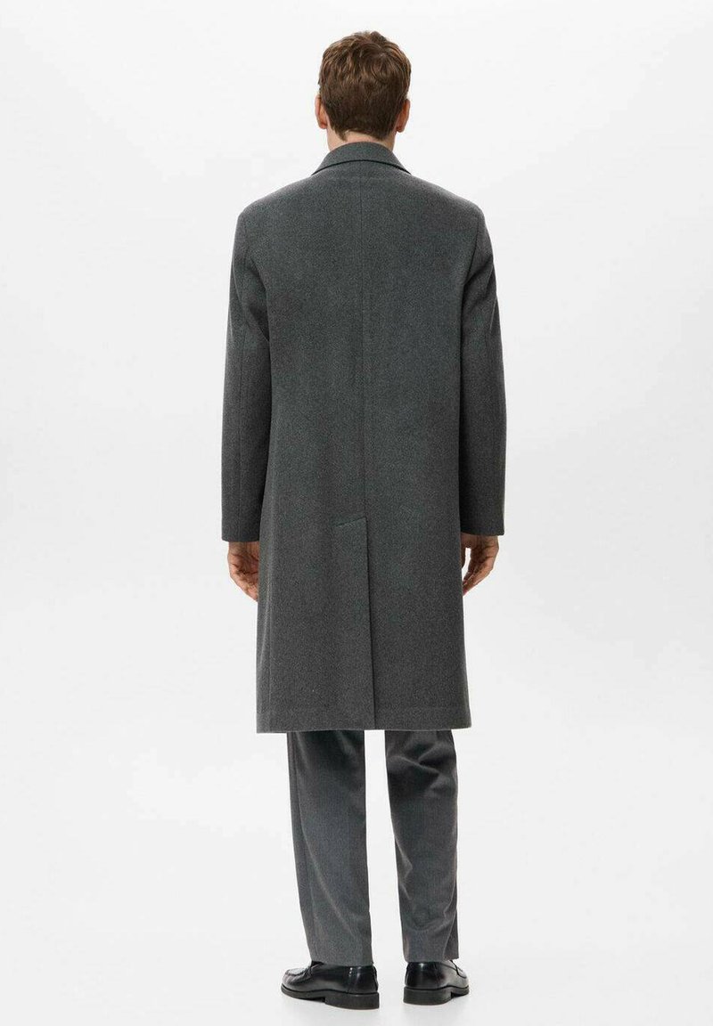 Lång grå ullkappa med rak snitt, enkel vent bak och utan synliga stängningar. Bärs över grå byxor och svarta skor.