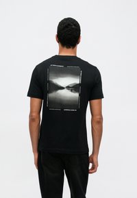 Czarna bawełniana koszulka z prostokątnym nadrukiem fotograficznym na plecach, przedstawiającym jezioro i góry w odcieniach szarości, z tekstowymi akcentami.