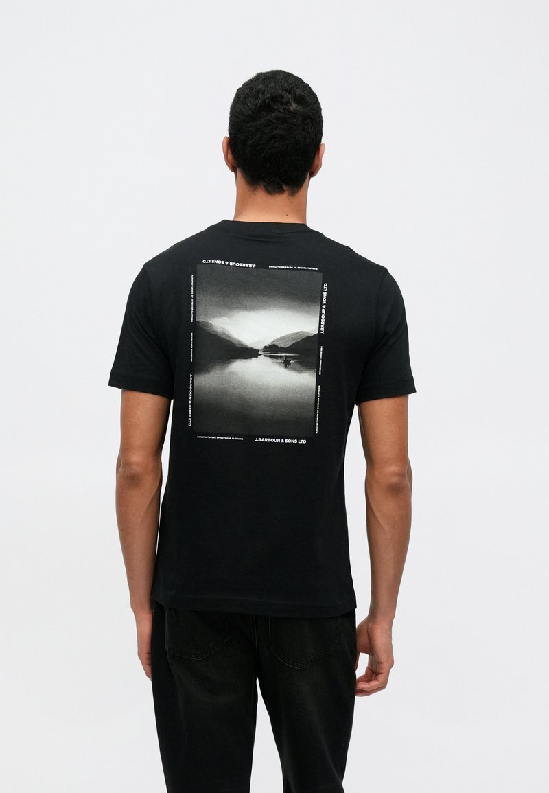 Czarna bawełniana koszulka z prostokątnym nadrukiem fotograficznym na plecach, przedstawiającym jezioro i góry w odcieniach szarości, z tekstowymi akcentami.