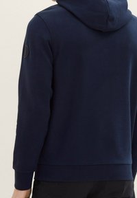 Mörkblå hoodie med en mjuk textur, med en dragsko i huvan, ribbade muddarna och en rund logotypapplikation på vänster axel.