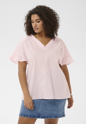 Vrouw met krullend haar, gekleed in een lichtroze blouse met ruches en V-hals en een blauwe spijkerrok, staand tegen een effen achtergrond.