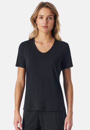 Frau mit schulterlangen blonden Haaren, die ein schlichtes schwarzes kurzärmliges V-Ausschnitt-Shirt und schwarze Hosen trägt und vor einem weißen Hintergrund steht.