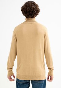 Suéter de cuello alto beige con puños y dobladillo acanalados, con una textura suave y lisa y un corte holgado, usado sobre jeans azules.