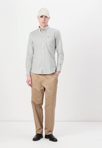 Chemise à carreaux grise et blanche à manches longues, assortie à un pantalon beige clair. La tenue est complétée par une casquette de couleur claire.