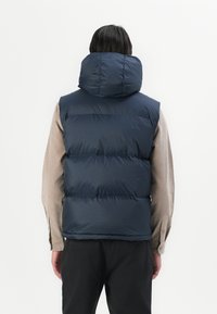 Navy blue puffer vest with a high, rounded collar, featuring horizontal stitching and a smooth, shiny texture. Worn over a light shirt.