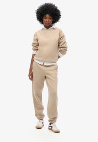 Beige bomulds sweatshirt og matchende joggers med manchetter ved anklerne. Hvid skjorte med krave under. Sneakers med sorte og brune accenter.