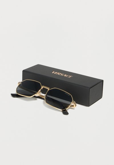 Γυαλιά ηλίου με μαύρους φακούς και χρυσά γεωμετρικά πλαίσια, τοποθετημένα μπροστά από μια μαύρη ορθογώνια θήκη Versace σε λευκή επιφάνεια.
