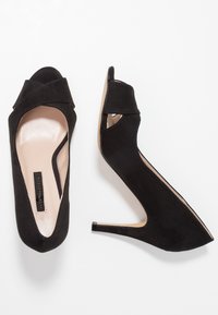 Černé semišové lodičky na vysokém podpatku s odhaleným prstem a zkříženým designem v přední části. Mají zúžený podpatek a hladkou koženou stélku.