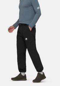 Zwarte broek met riemlussen, voorzakken en een wit logo op de linkerdij. Draagt een lichtblauw long-sleeve shirt en groene schoenen.