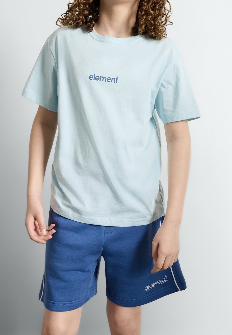 Jeune portant un t-shirt bleu clair « element » et un short bleu foncé avec le logo « element », debout devant un fond clair uni.